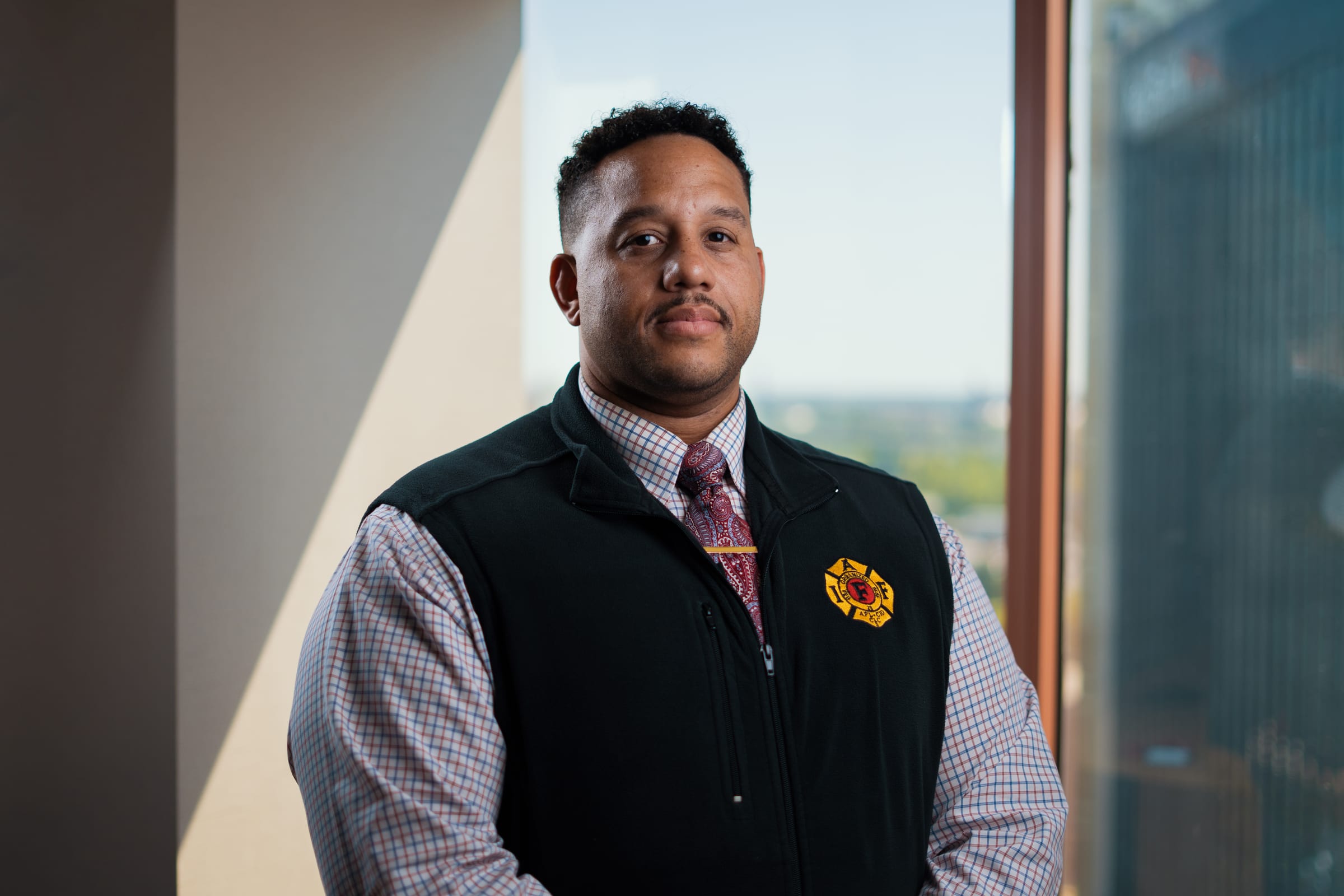 A professional portrait of Joshua Archer, a man with short dark hair and a mustache, standing and looking forward with a neutral expression. He is wearing a plaid button-down shirt, a patterned red tie secured with a tie clip, and a dark fleece vest featuring an International Association of Fire Fighters (IAFF) emblem on the chest. His hands are clasped in front of him, and he is positioned indoors near a window with an out-of-focus background.