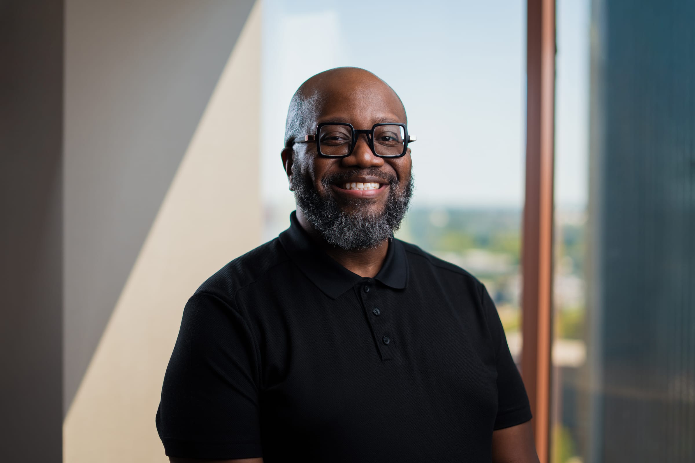 A professional portrait of Randall Parker III, a bald man with a full graying beard and distinctive black-framed glasses, smiling warmly at the camera. He is wearing a black polo shirt, and is positioned indoors near a window with an out-of-focus background.
