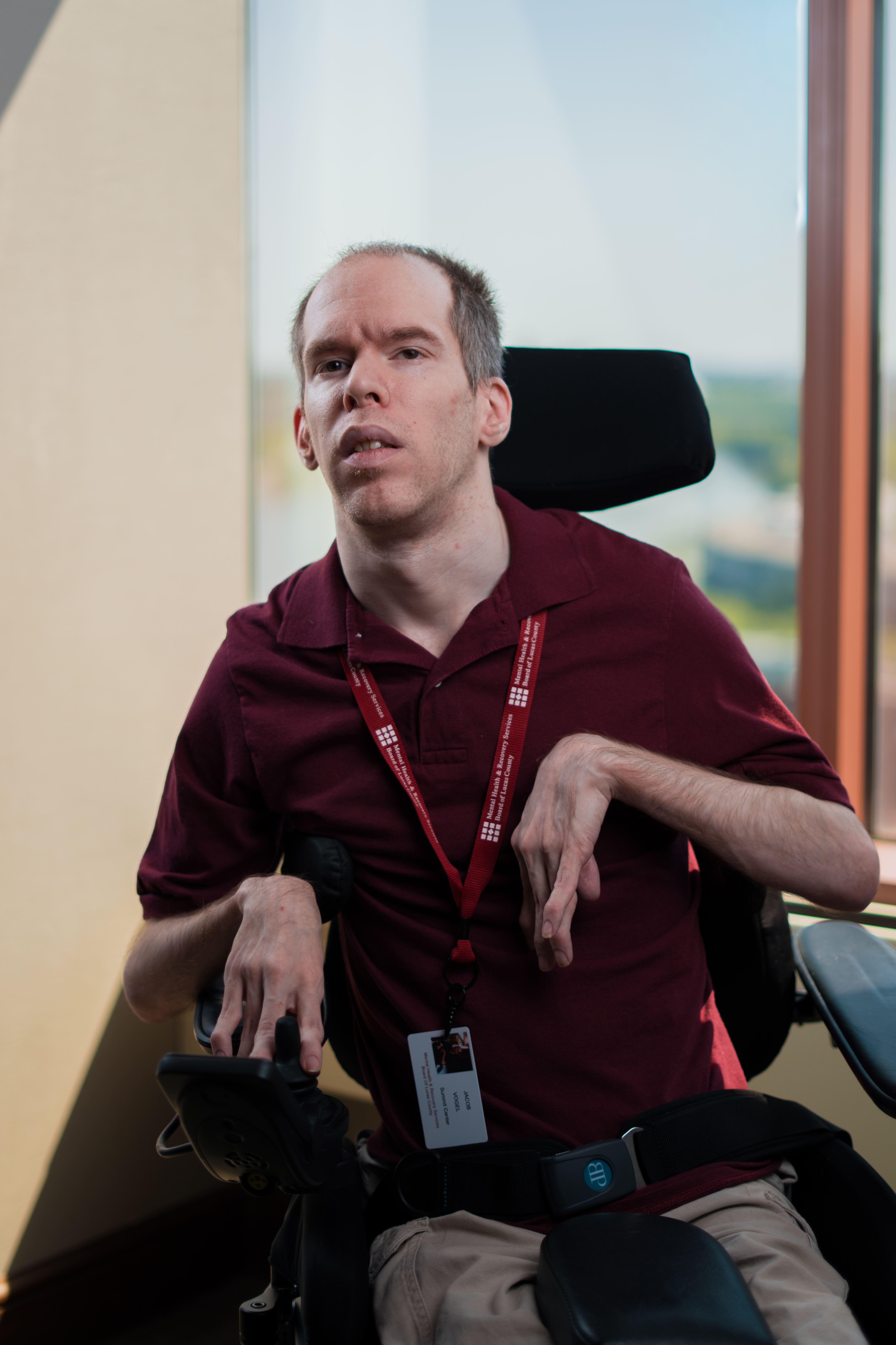 A professional portrait of Jacob Vogel, a man seated in a power wheelchair indoors near a window with an out-of-focus background. He is wearing a maroon polo shirt and a red lanyard with an ID badge from the Mental Health & Recovery Services Board of Lucas County. He has short hair and is looking forward.